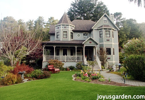 a large green/grey house sits set back with an artificial lawn & a beautiful garden