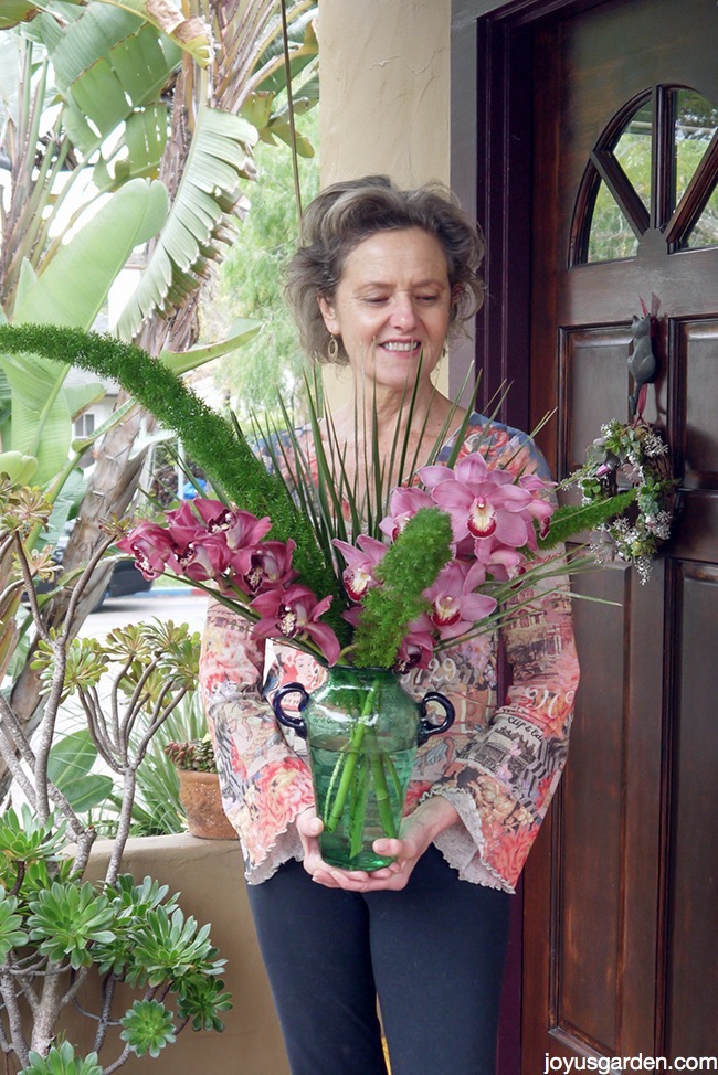 nell foster smiling and holding a foxtail fern flower arrangement in front of her front door