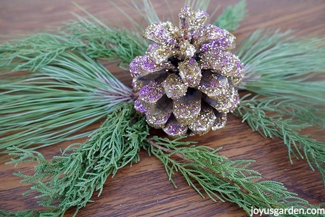 a pine cone glittered with gold & purple glitters sits on evergreen branches