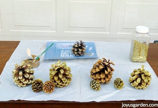 gold glittered pine cones, a jar of gold glitter, a saucer with gold paint & 2 brushes & a flexible cutting board sit on a piece of paper on a table