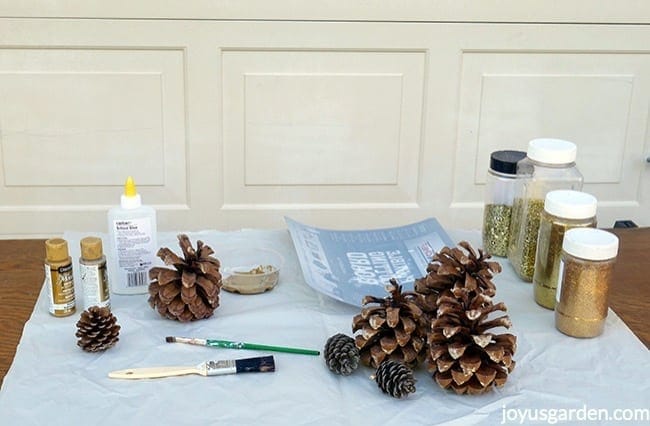 Pine cones, 4 types of gold glitters, school glue, metallic paints, 2 brushes & a flexible cutting board sit on a work table