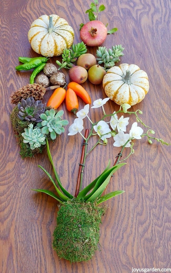 all the ingredients for a fall table decoration including a white orchid, small pumpkins, fruits & succulents lay on top of a table