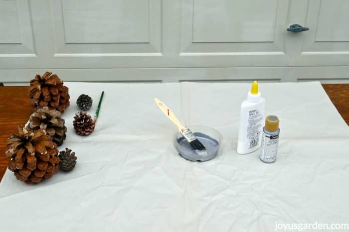 2 sizes of pine cones, a saucer with paint & a brush, school glue & a jar of silver metallic paint sit on a work table