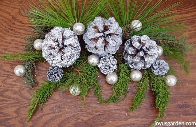 A decoration of silver glittered pine cones with pearlescent balls & fresh evergreen branches sit on a table.