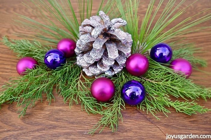 Close up of a silver & pink glittered pine cone adorned with hot pink & purple balls & fresh evergreen branches 