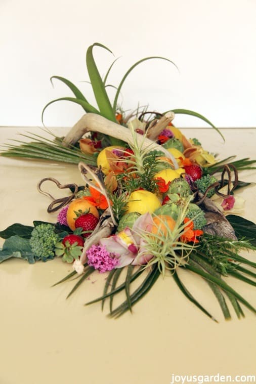 a long arrangement with driftwood, fruits, vegetables, air plants, flowers, an agave & foliages sit on a pale yellow table