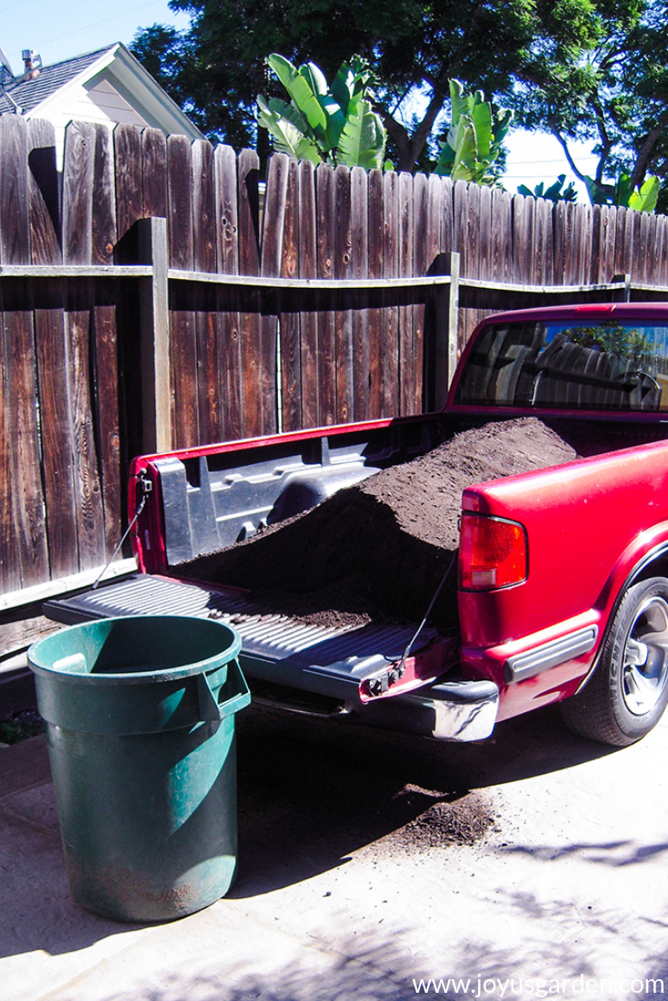 the back of an empty green barrel sits next to a red truck filled with soil amendments