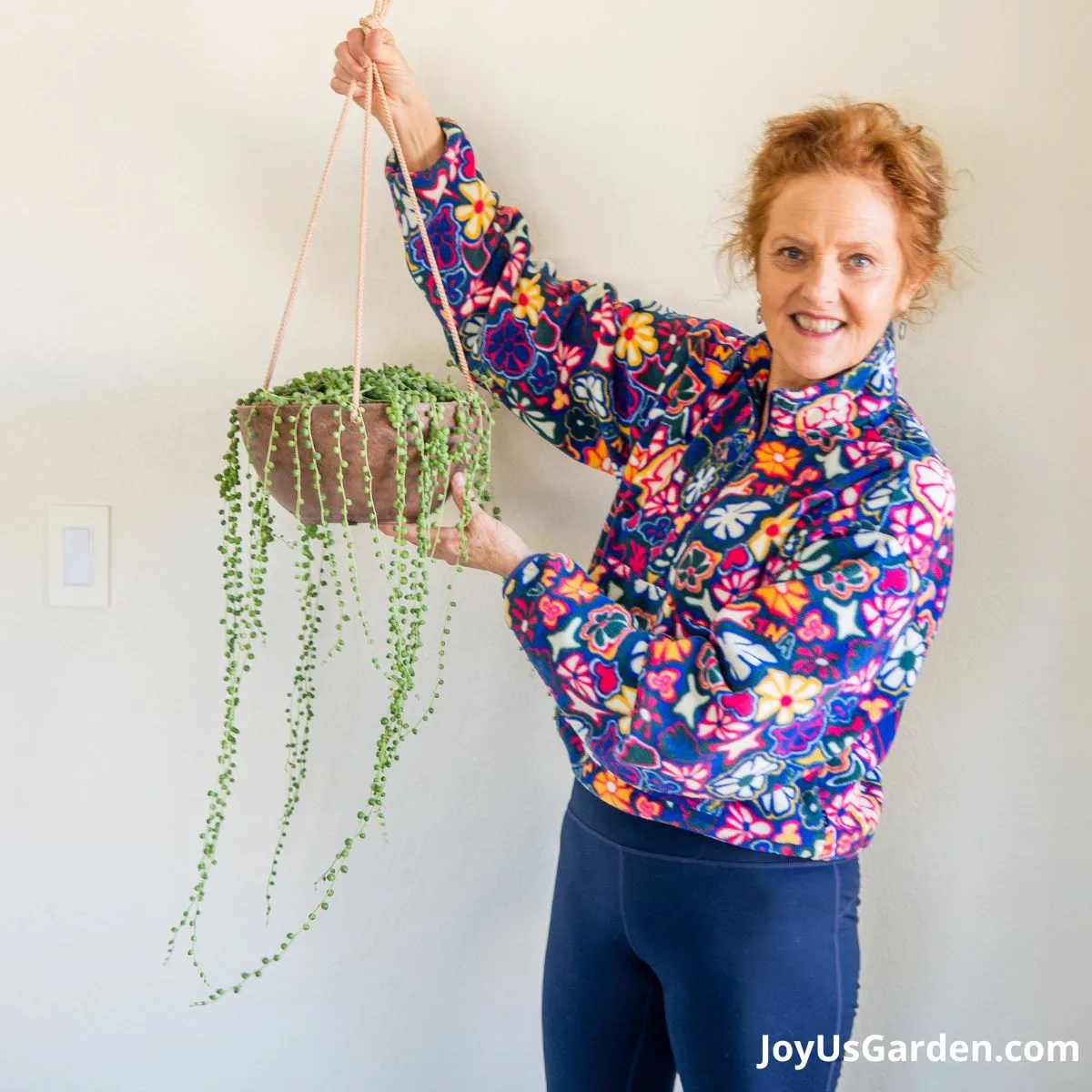 nell foster incolorful floral sweater standing indoors holding a string of pearls plant
