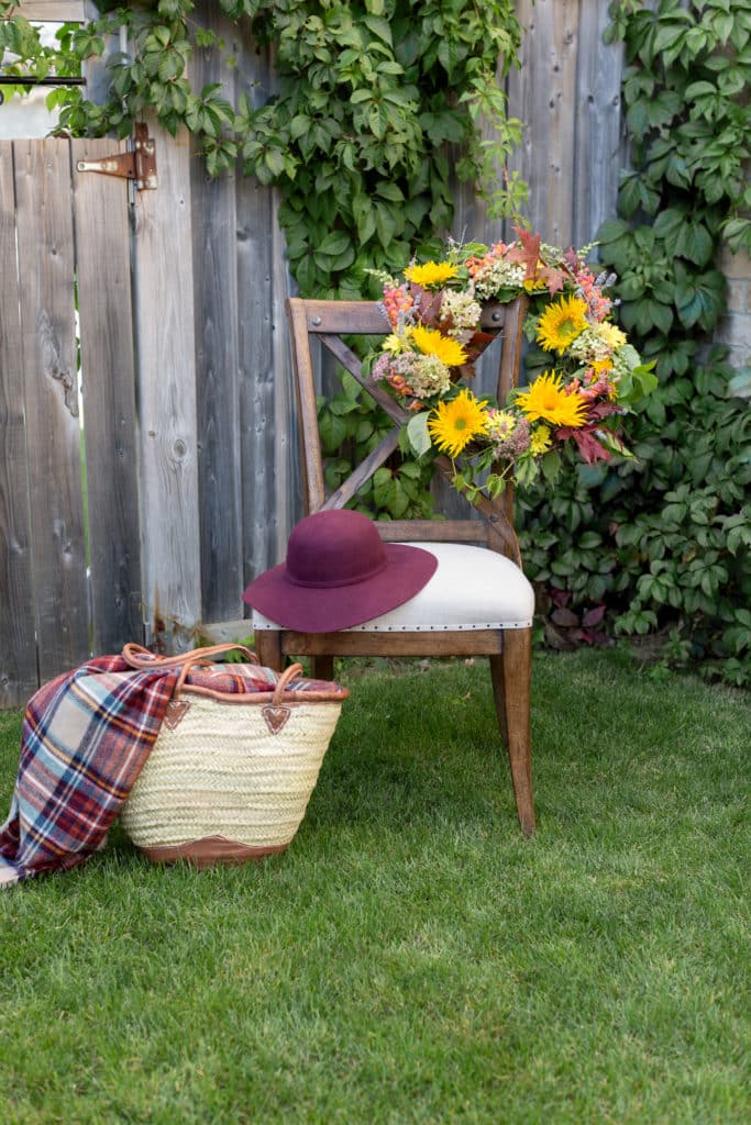 a colorful fresh floral fall wreath hangs off a chair. a red hat & a basket sit next to the wreath