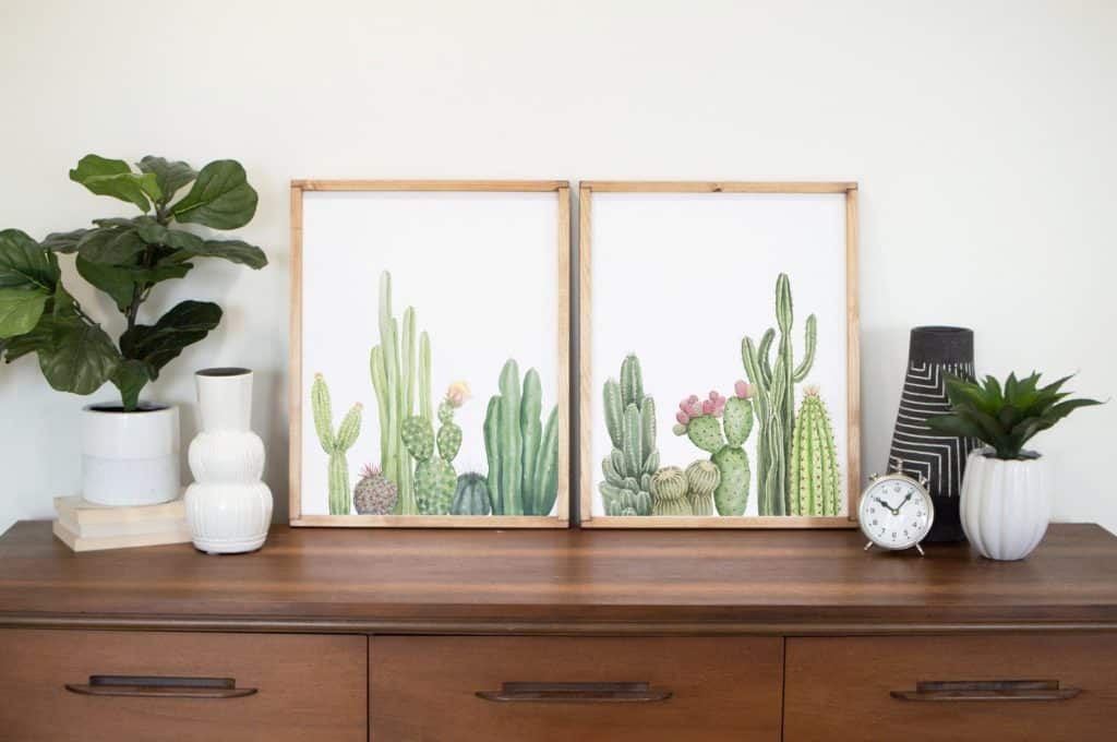 two prints of green cacti with pink flowers on a dresser with white vases and plants