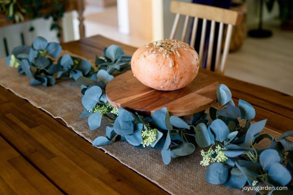 a small peanut pumpkin sits on a wooden cake stand on top of a burlap table runner with eucalyptus garland running down the table