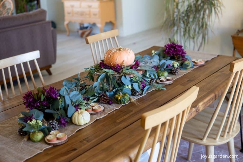 a thanksgiving centerpiece diy consisting of eucalyptus garland, gourds, pinecones, tea candles, a peanut pumpkin on a cake stand, wheat heads & burgundy mums