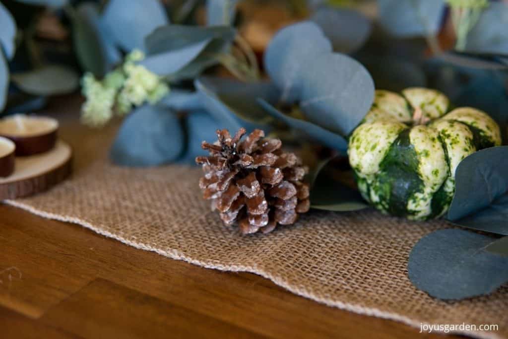 close up of a thanksgiving centerpiece with a burlap table runner a glittered pinecone a green & white gourd tea candles & eucalyptus garland