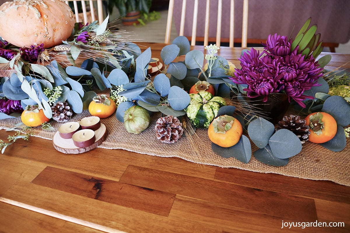 close up of a fall table decorated with a burlap table runner, eucalyptus garland, burgundy mums, wheat heads, tomatillos, gourds, pinecones, tea candles, & persimmons