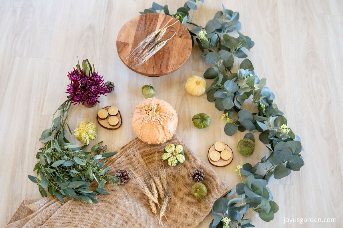 materials to be used to make DIY thanksgiving centerpiece: eucalyptus garland, pumpkins, gourds, wood blocks, seeded eucalyptus, cake stand, wheat bundle, pinecones, candles, and mums