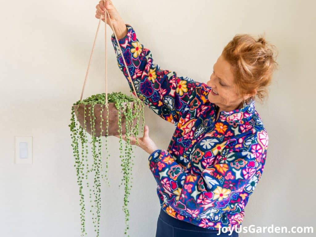 nell foster wearing a floral sweater holding hanging planter bowl with string of pearls trails hanging over the sides 