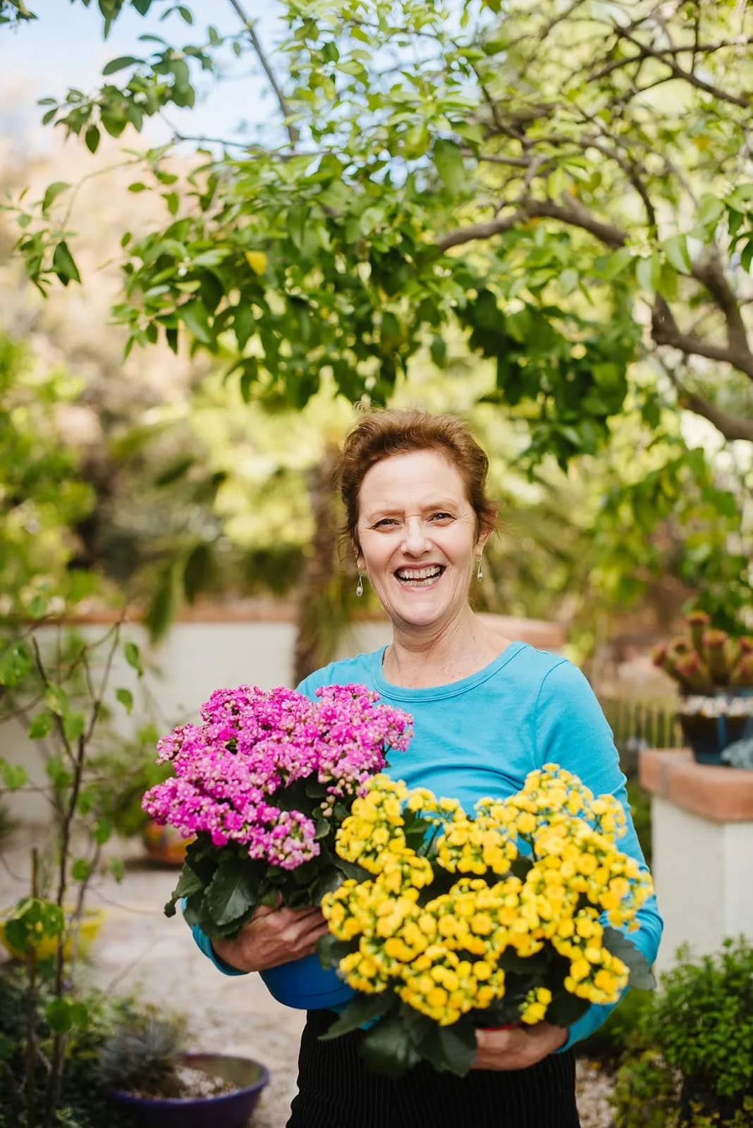 Now Foster stands outside holding two kalanchoes, in pink and yellow in a turquoise top.