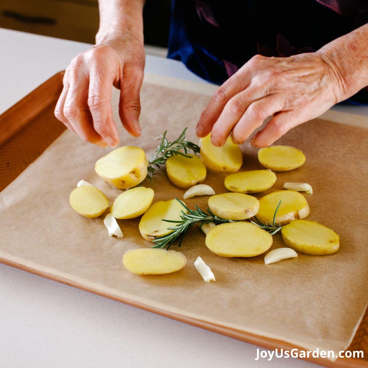 woman is shown cooking using rosemary, on the cookie sheet is rosemary sprigs, potatoes, and and garlic