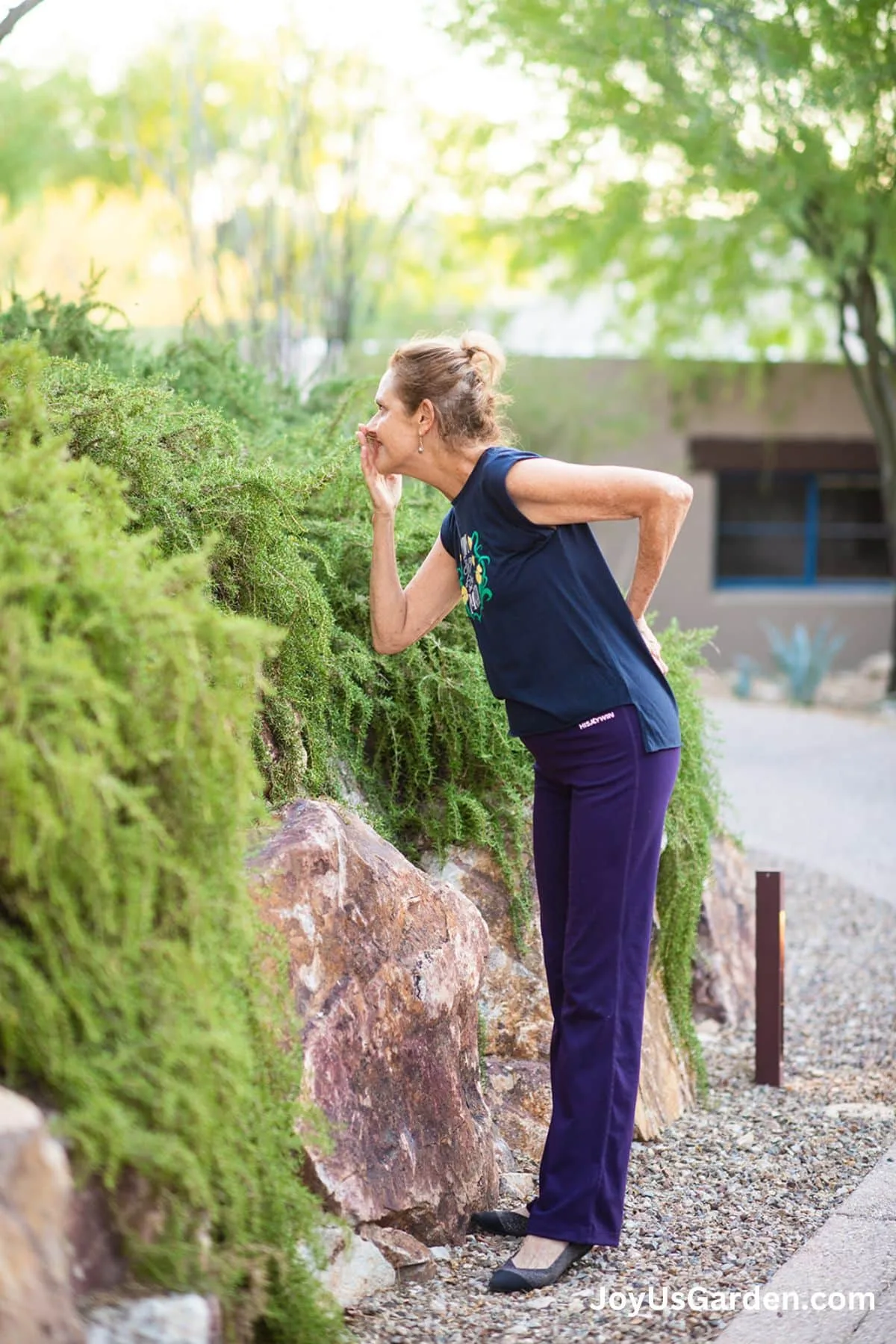 Nell Foster wearing a blue blouse and purple pants leans in and smells rosemary fragrant leaves