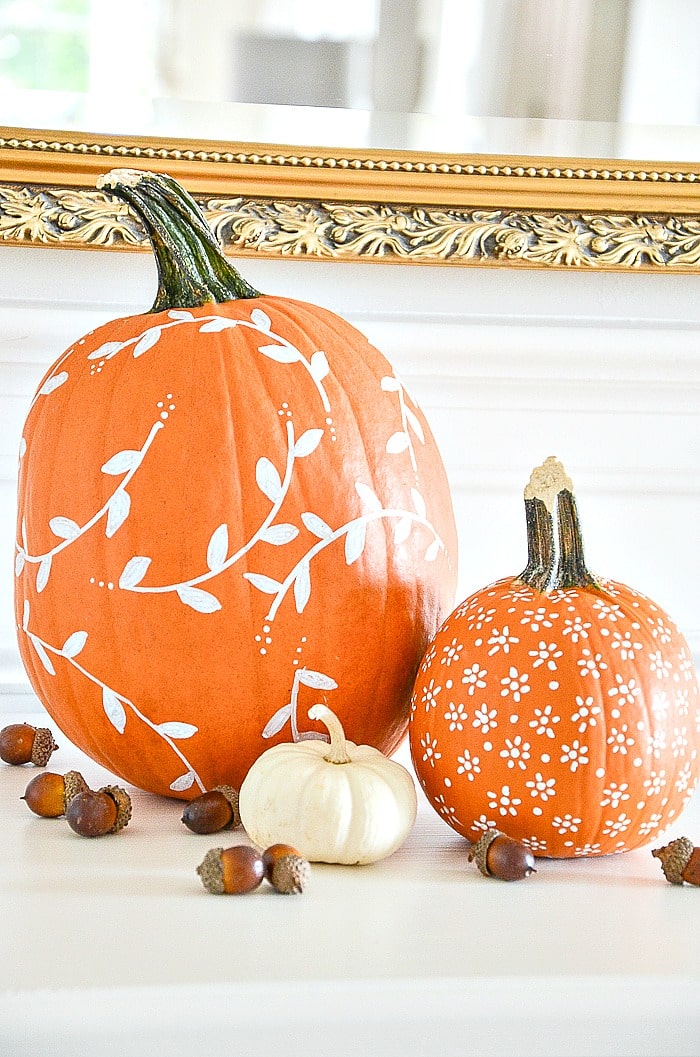 2 orange pumpkins painted with a white design sit on a table with a white mini pumpkin & acorns.