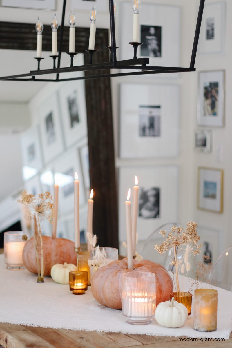 Orange/pink pumpkins with candles line a table with white mini pumpkins, dried stems, & candles in glass votive cups. 