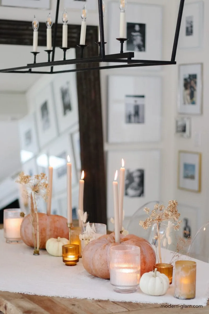 Orange/pink pumpkins with candles line a table with white mini pumpkins, dried stems, & candles in glass votive cups.