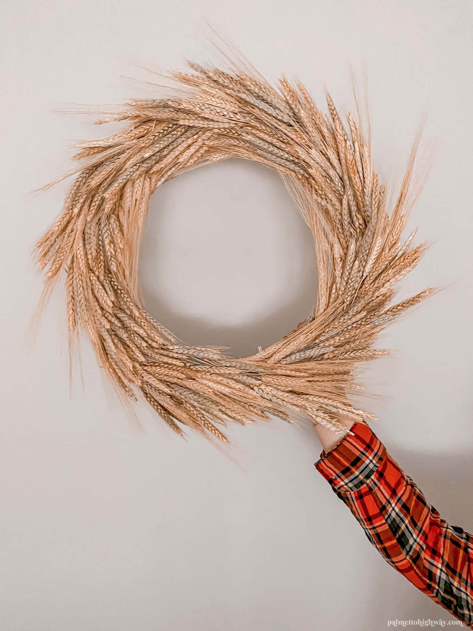 A hand holds a fall wreath created from wheat against a wall.