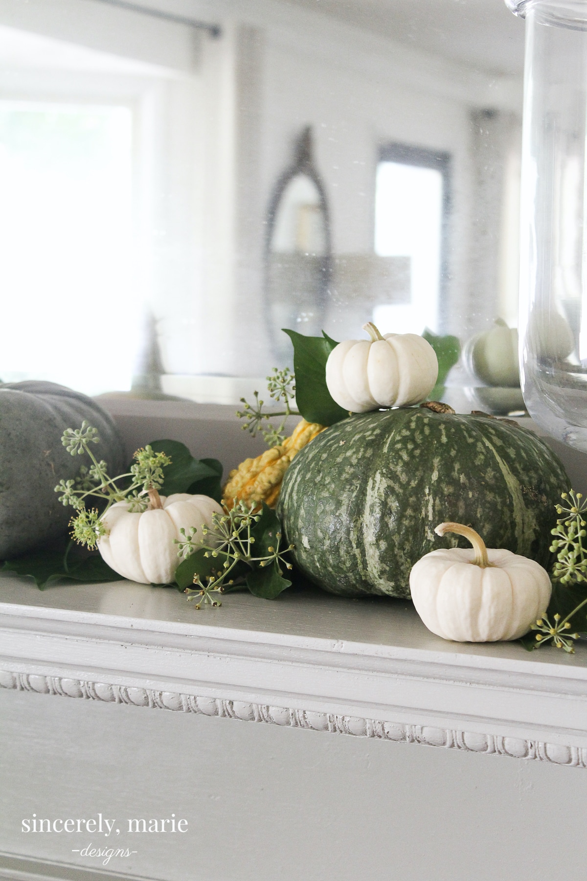 A fireplace mantel adorned with large green pumpkins, small white pumpkins, a yellow gourd, & seeded eucalyptus.