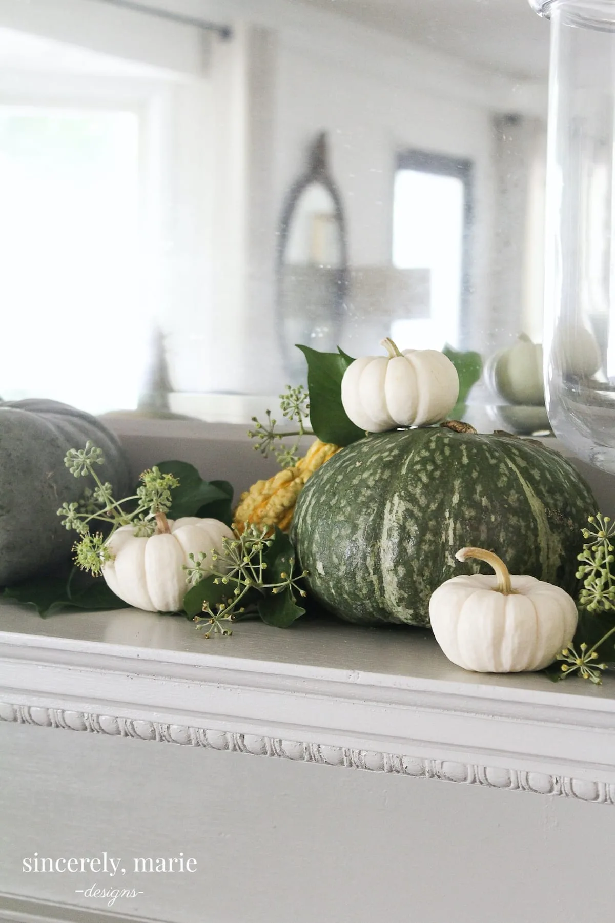 A fireplace mantel adorned with large green pumpkins, small white pumpkins, a yellow gourd, & seeded eucalyptus.