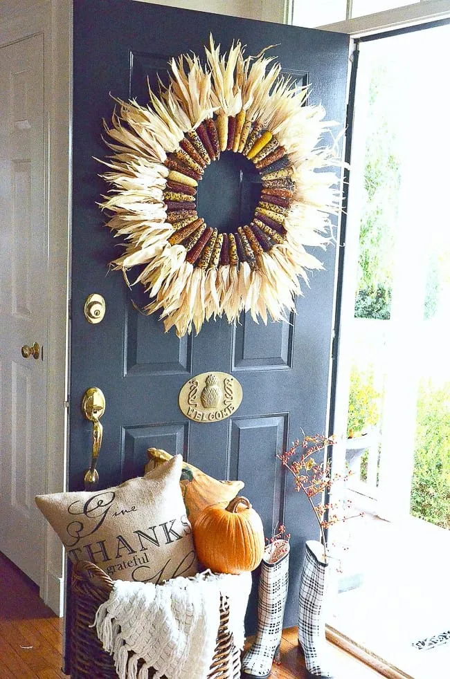 A fall wreath made from multi-colored mini corns hangs on a dark blue door. A basket sits at the base.