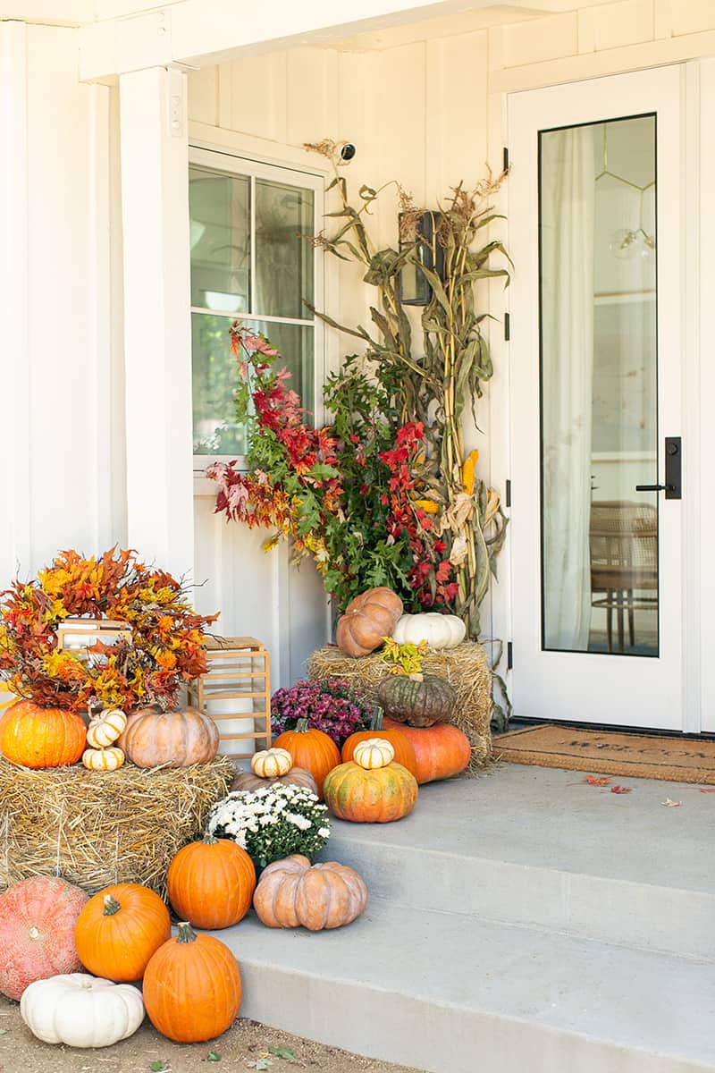A front porch decorated beautifully for fall with corn stalks, foliage stems, hay bales, an assortment of pumpkins, & mums.