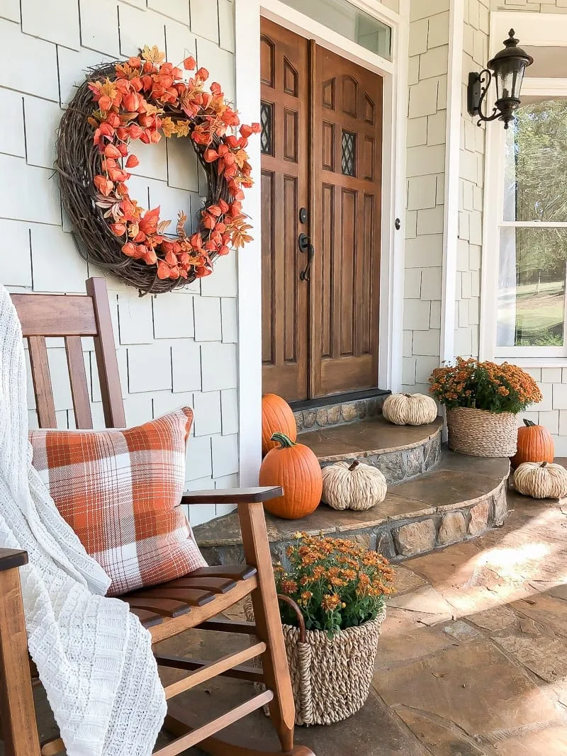 A front entry way decorated for fall with orange & beige pumpkins, 2 baskets with orange mums, a rocking chairs, & a large grapevine wreath with chinese lanterns. 