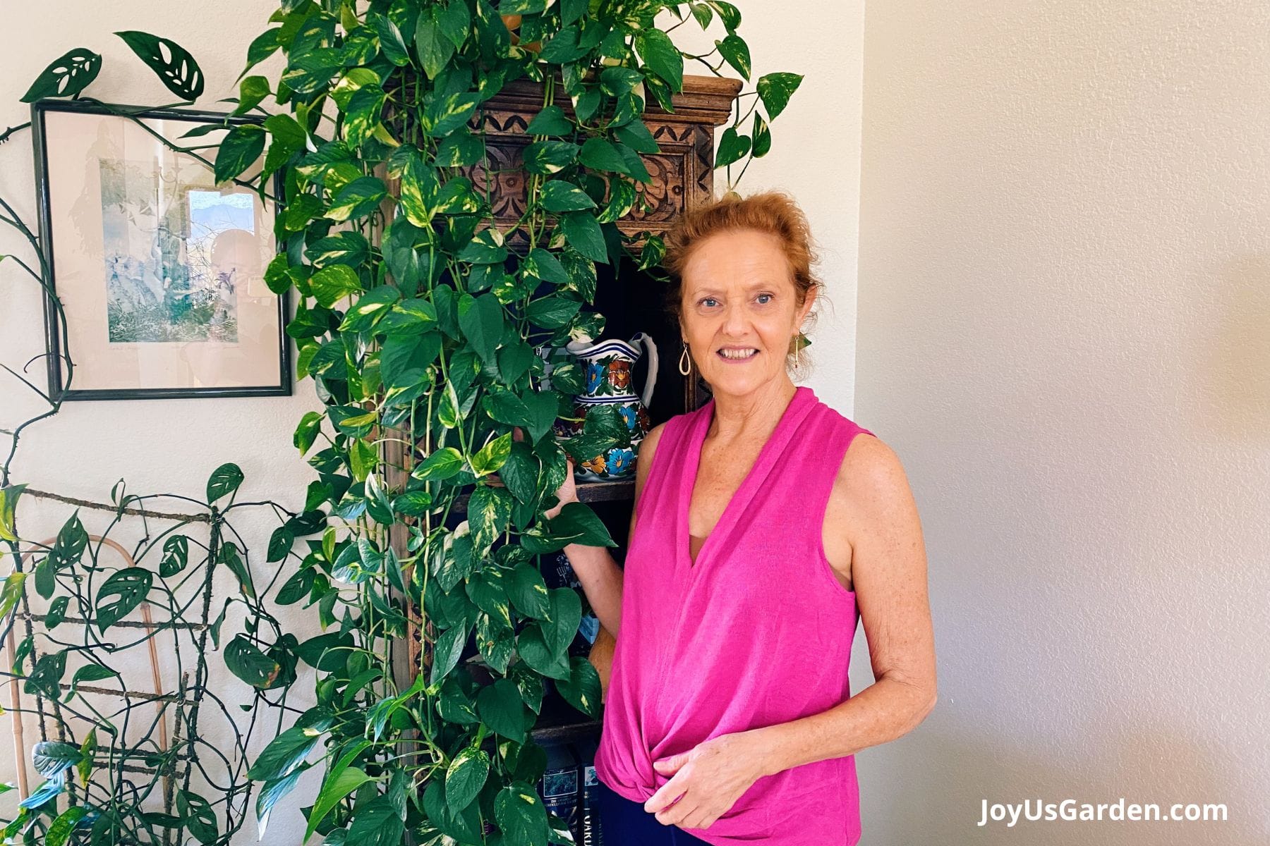 Nell Foster in a pink blouse stands next to her golden pothos on top a bookshelf.